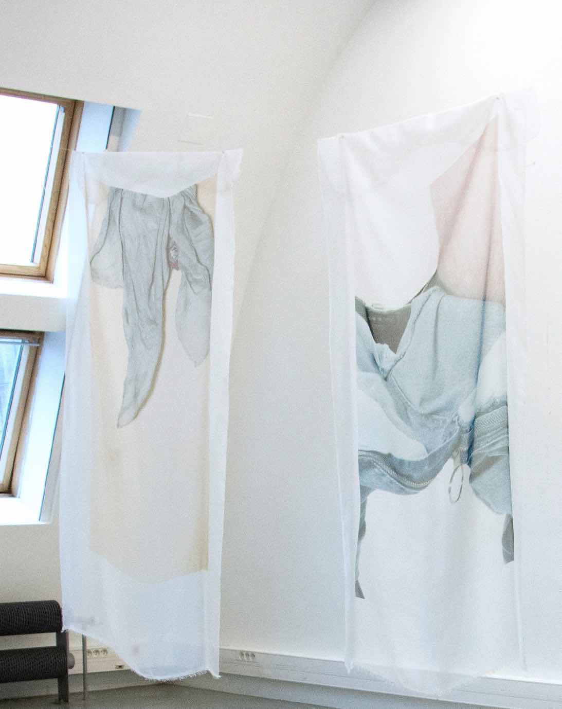 Two polyester silks hanging from the ceiling. Printed onto the silk are photographs of a closeup view of a leg with the jeans pants stripped down to the foot and another photograph zoomed up to a male chest 
            with a blue scarf around the neck. The nippel and the nipplehair is barely visbile.